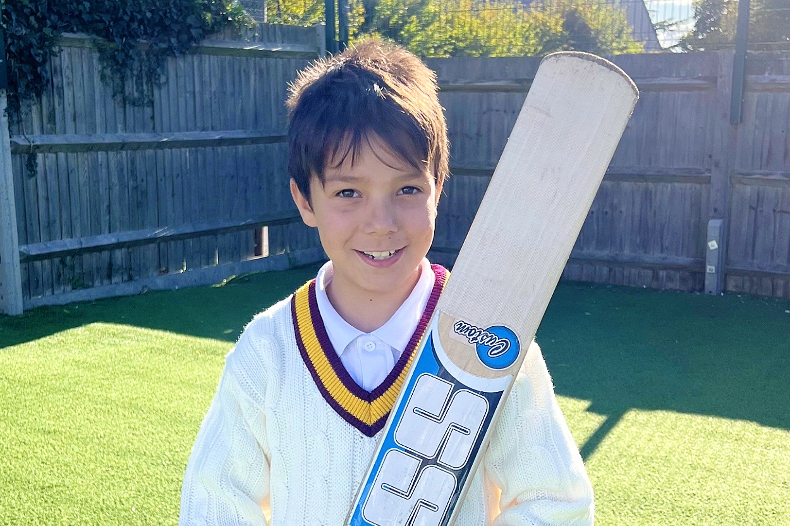 A young pupil in cricket whites with a burgundy and gold trimmed jumper, smiling and holding a cricket bat
