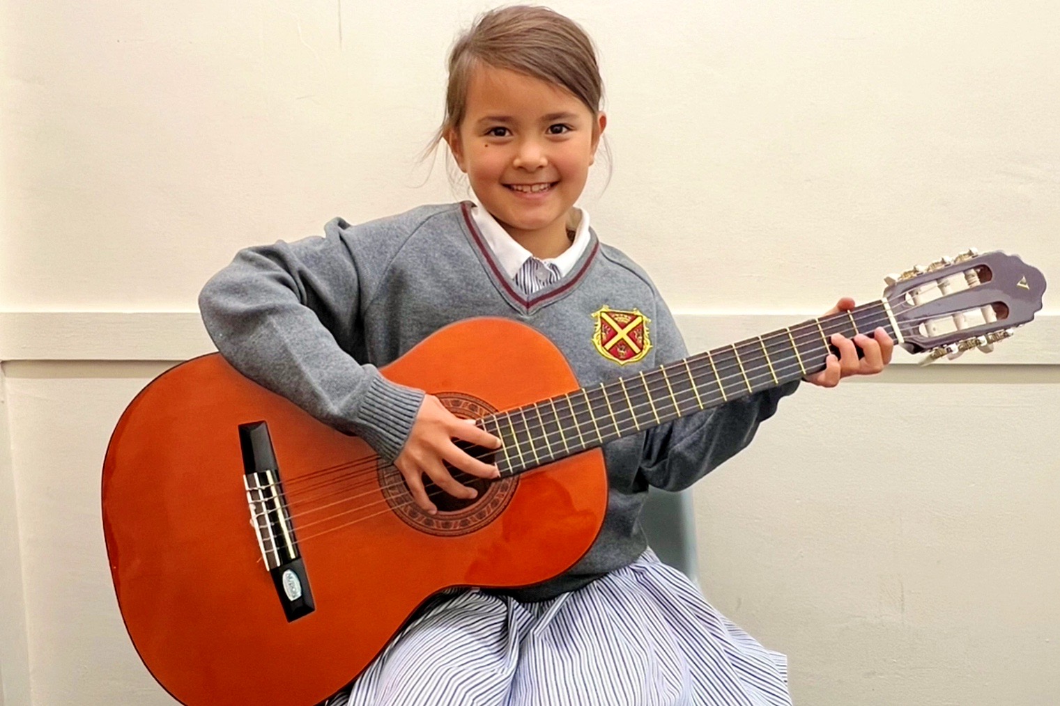 A young pupil in a Queen's Square jumper smiling and holding a classical guitar